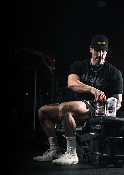 A muscular man in a gym setting wearing a black 'ANIMAL' t-shirt black shorts and white sneakers sitting on a bench with shaker bottles.